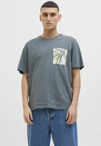 Camiseta gris de manga corta con una textura especial, que presenta un bolsillo en la parte delantera de color blanco decorado con un diseño de hojas amarillas y verdes.