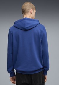 Blauer Pullover mit Kapuze und Kängurutasche. Glatte Textur, lange Ärmel und eine Kapuze mit Kordelzug. Schwarze Hose mit sichtbaren Taschen.