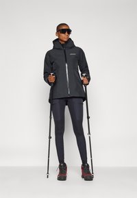 Black waterproof jacket with hood, silver zipper, and logo. Worn with dark leggings and red-accented hiking boots, holding trekking poles.