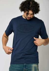 T-shirt à manches courtes bleu marine en coton doux, avec un logo "Kaporal" en relief et un design col rond. Coupe décontractée avec une texture lisse.