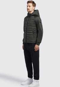Dunkelgrüne Pufferjacke mit Kapuze, gesteppter Optik, seitlichen Reißverschlusstaschen und gerippten Bündchen, kombiniert mit schwarzen Sweatpants und weißen Sneakers.