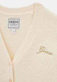 Cardigan crema con scollo a V, bordi a coste e superficie testurizzata. Presenta "Guess" ricamato in oro sul lato sinistro.