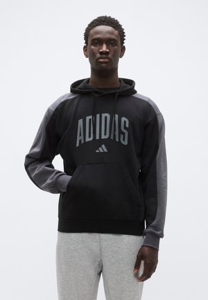 Sweat à capuche noir et gris avec grand logo "ADIDAS", poche kangourou, capuche à cordon et manches contrastantes. Porté avec un pantalon de survêtement gris.