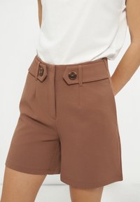 Femme portant un short marron taille haute avec de grandes poches à rabat boutonnées, accompagnée d'un haut blanc sans manches rentré à l’intérieur.