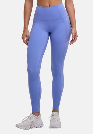 Vrouw die lavendelkleurige leggings met hoge taille en zijzakken draagt, samen met witte sportschoenen, staand tegen een effen achtergrond.