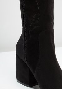 Botín de ante negro con tacón cuadrado, equipado con cremallera lateral y diseño de hojas en relieve en el cañón. Textura lisa en toda la superficie.