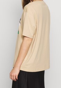 Beige bomulls t-shirt med kort ärm, lös passform, rund halsringning, med en grafik på framsidan och en rak fåll.