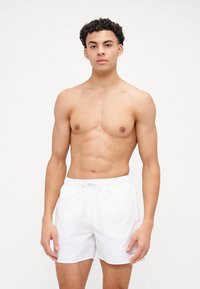 Shorts de bain blancs avec une taille élastique et un cordon de serrage, arborant un logo sur le côté inférieur droit. Texture lisse, coupe décontractée.