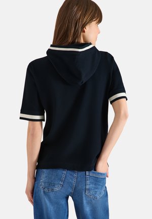 Femme portant un haut à capuche bleu marine à manches courtes avec un liseré blanc, assorti à un jean en denim bleu, vue de dos sur un fond uni.