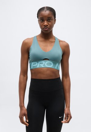 Mujer atlética con sujetador deportivo azul verdoso y leggings negros de talle alto, de pie contra un fondo blanco liso.