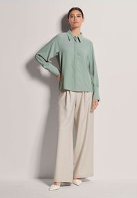 Hellgrünbluse mit Kragen, langen Ärmeln und Knopfmanschetten. Kombiniert mit cremefarbenen, weiten Hosen und spitzen weißen Absatzschuhen.