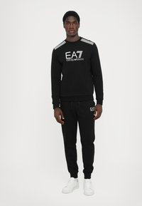 Black sweatshirt and joggers with logo detailing and stripe accents, made of a soft fabric. Paired with white sneakers.