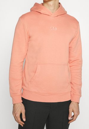 Homme portant un sweat à capuche GAP rose saumon avec poche avant et pantalon noir, visible du cou aux hanches sur un fond uni.