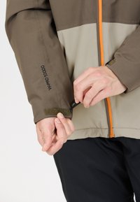 Brun och beige jacka med en orange dragkedja och justerbart handledsband. Har strukturerad tyg och en logotyp på ärmen.