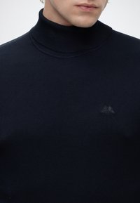 Mann trägt einen schwarzen Rollkragenpullover mit kleinem gesticktem Logo auf der linken Brust, gezeigt werden nur das untere Gesicht und der Hals.