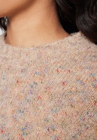 Maglione beige texturizzato con scollo rotondo, caratterizzato da una varietà di macchie colorate in tutto il tessuto, creando un aspetto maculato.