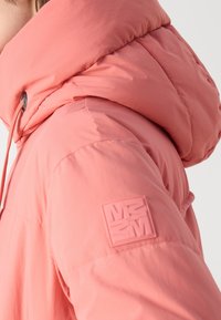 Personne portant une veste matelassée rose avec une grande capuche et un patch logo embossé sur le haut de la manche.