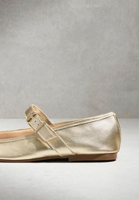 Zlatá metalická plochá obuv Mary Jane s nastavitelným přezkovým páskem a nízkou hnědou podrážkou na světle šedém povrchu.