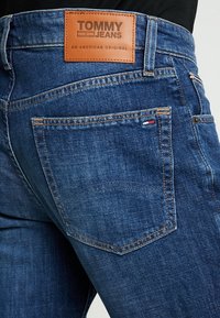 Jeans de mezclilla en azul oscuro con un parche de cuero en la parte posterior grabado con "TOMMY JEANS." Presenta un bolsillo trasero y costuras en contraste.
