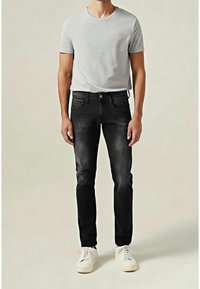 Homme portant un t-shirt gris clair à manches courtes, un jean noir slim-fit et des baskets blanches, debout devant un fond neutre.