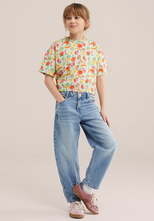 Dijete stoji s rukama u džepovima, nosi šarenu košulju s uzorkom voća, svijetloplave traperice, bijele čarape i ružičaste tenisice.