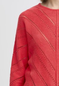Korallenfarbener Strickcardigan mit einem strukturierten Muster und diagonalen Ausschnitten. Das Design hat einen runden Halsausschnitt und sanft fallende Ärmel.