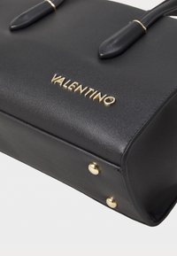 Černá kožená kabelka s hladkým povrchem, zdobená kováním v barvě zlata a vyraženým logem "VALENTINO" na přední straně, kulatým tvarem a pevnými rukojetmi.