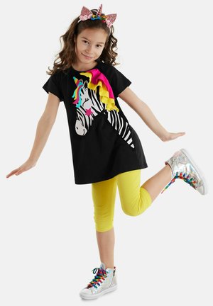 Schwarzes T-Shirt mit einem Zebra mit einem Regenbogenmähne, kombiniert mit gelben Capri-Leggings und schimmernden High-Top-Sneakern mit bunten Schnürsenkeln.