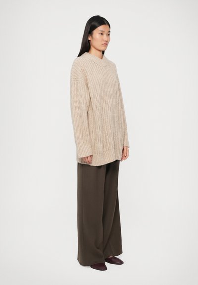 Beige gestrickter Pullover mit V-Ausschnitt, locker sitzend und mit Rippenstruktur, kombiniert mit olivfarbenen weit geschnittenen Hosen und dunkelroten Schuhen.