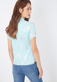 Camisa de manga corta azul claro con grandes lunares plateados, que presenta un escote redondeado y una abertura lateral para mayor facilidad de movimiento.