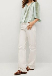 Blusa leggera verde chiaro, trasparente, con scollo a V e maniche morbide, abbinata a pantaloni bianchi aderenti e sandali marroni con cinturini.