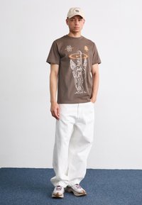 Brun t-skjorte med grafisk trykk, med et stilisert engeldesign. Kombinert med løse hvite denimbukser og nøytrale sneakers.