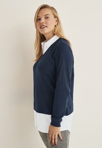 Marinblå v-ringad tröja lager på lager över en vit skjorta med knappar, med långa ärmar och en något avslappnad passform. Texturerade grå rutiga byxor.