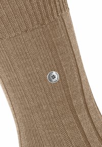 Calzino lavorato a maglia beige con texture a coste, una cima arrotondata e un logo rotondo argentato attaccato di lato.