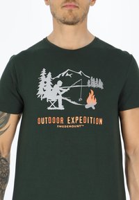 Mørkegrøn bomulds T-shirt med en fiskescene, der viser en person, træer, bjerge og et lejrbål i hvid og orange grafisk tryk.