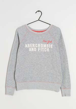 Sudadera gris con mangas raglán y puños acanalados. Presenta el texto "Abercrombie and Fitch" y "New York" en blanco y rojo. Cuelga de un gancho negro.