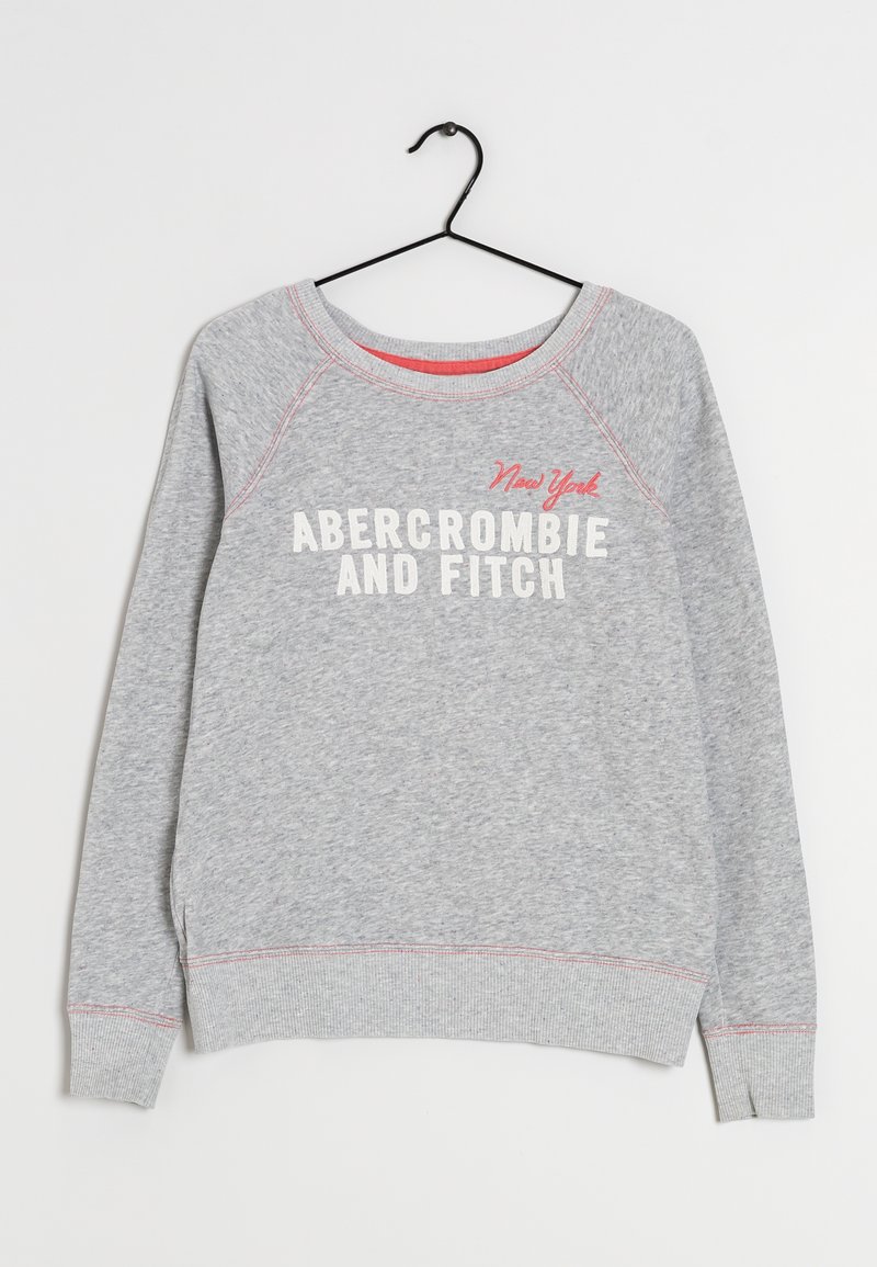 Sudadera gris con mangas raglán y puños acanalados. Presenta el texto "Abercrombie and Fitch" y "New York" en blanco y rojo. Cuelga de un gancho negro.