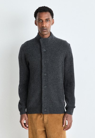Cardigan en maille gris foncé avec un col haut, un texture côtelée et une fermeture à boutons. Manches longues et coupe structurée. Porté par-dessus un pantalon en velours côtelé beige.