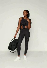 Črna športna kratka majica z detajlom pentlje v kombinaciji z visokim pasom črnih pajkic. Obleko dopolnjujejo beli supergi in črna potovalna torba.