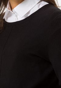 Černý svetr s kulatým výstřihem, vyrobený z měkkého materiálu. Na výstřihu vykukuje bílá košilová halenka. Hladká textura.