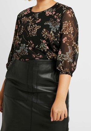 Femme portant un chemisier transparent noir à motif floral avec des manches 3/4 froncées et une jupe en cuir noire taille haute avec poches avant.