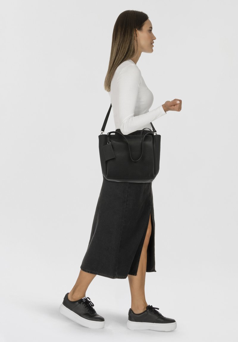 Sac fourre-tout en cuir noir avec une finition texturée, poignées supérieures doubles et une étiquette amovible. Le modèle porte un haut blanc et une jupe noire, marchant.