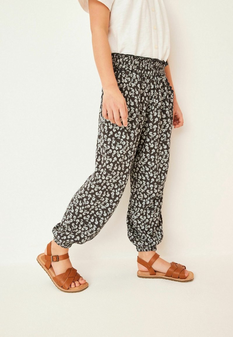 Personne portant un pantalon à taille élastique noir et blanc à imprimé floral et des sandales en cuir marron avec des brides à boucle.