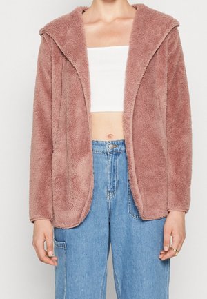 Person trägt eine staubig rosa flauschige Jacke über einem weißen, bauchfreien Top und hellblauen, hoch taillierten Jeans mit einem Ring an einem Finger.
