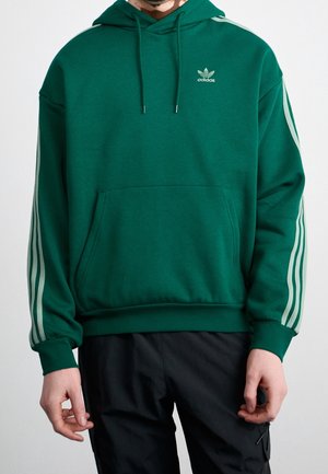 Person trägt einen grünen Adidas-Hoodie mit weißen Streifen an den Ärmeln und einer vorderen Tasche, kombiniert mit schwarzen Hosen.