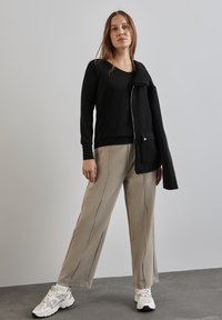 Top nero a maniche lunghe abbinato a pantaloni beige a gamba larga con cuciture laterali. Il modello indossa una giacca nera con mezzi zip e un colletto testurizzato.