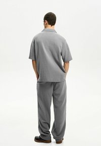Graues, strukturiertes Kurzarmhemd und passende locker sitzende Hose mit dezentem Crinkle-Muster und lässigem Design. Schwarze Schuhe vervollständigen den Look.