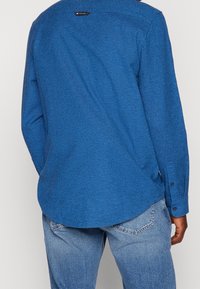 Blaues strukturiertes Langarmhemd mit abgerundetem Saum und geknöpften Manschetten, mit einem kleinen Etikett am hinteren Halsausschnitt. Kombiniert mit hellen Jeans.