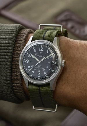 Timex MK1 automaatkäekell musta sihverplaadi, valgete numbrite ja rohelise kangasrihmaga, kantud vasakul randmel oliivsärgist kootud varruka kohal.