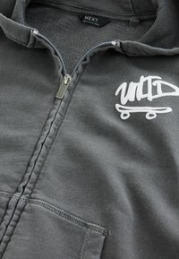 Grå zip-up hoodie lavet af blødt stof. Har en frontlomme, sølv lynlås og hvid grafisk logo med skateboarddesign.